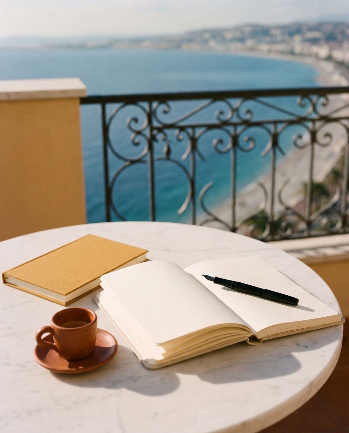Taccuino aperto, penna stilografica e caffè su un tavolino in terrazza affacciato sulla Baia degli Angeli